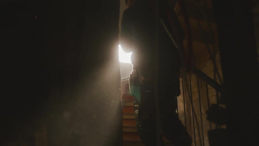 Man carries tubes up staircase from dim room as sun shines in brightly