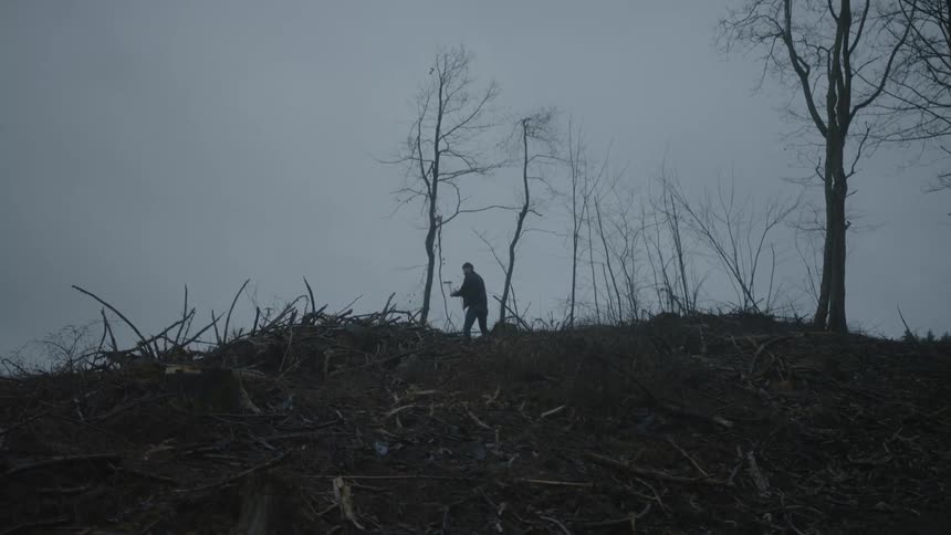 A man is standing on top of a hill swinging an axe high over his head as chops down a tree
