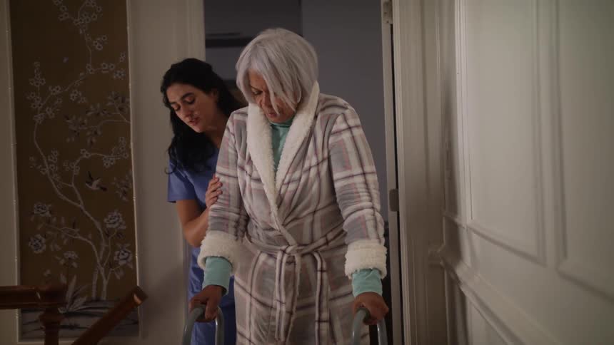 A healthcare worker helps a woman with a walker go down a hall