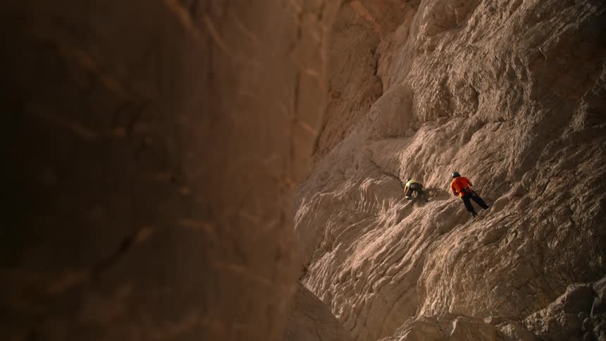 Two people rock climbing up a cliff one wearing orange and one wearing yellow