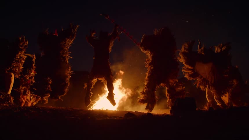 A group of people dressed in costumes jumping around a bonfire on the beach at night