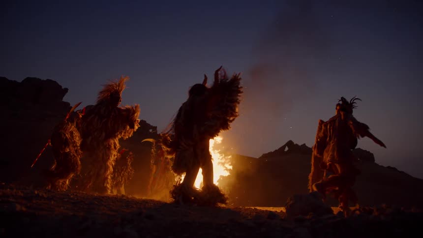 People dressed in costumes and masks dancing around a bonfire at night on the beach