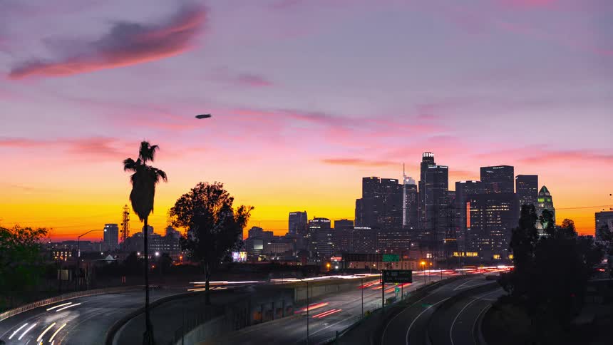 Towers and skyscrapers stand tall on the city skyline as traffic moves on the freeway and a blimp flies overhead