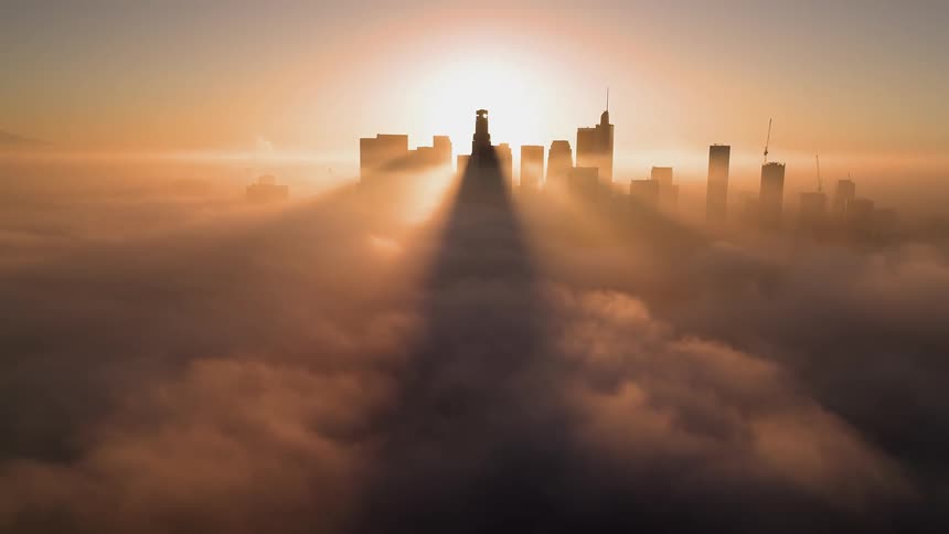 The sun shines brightly behind the city skyline and glowing rays illuminate the clouds as they move across the sky