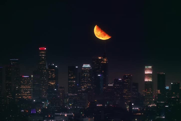 A time lapse of a half moon rising over a city lit up at night with tall buildings