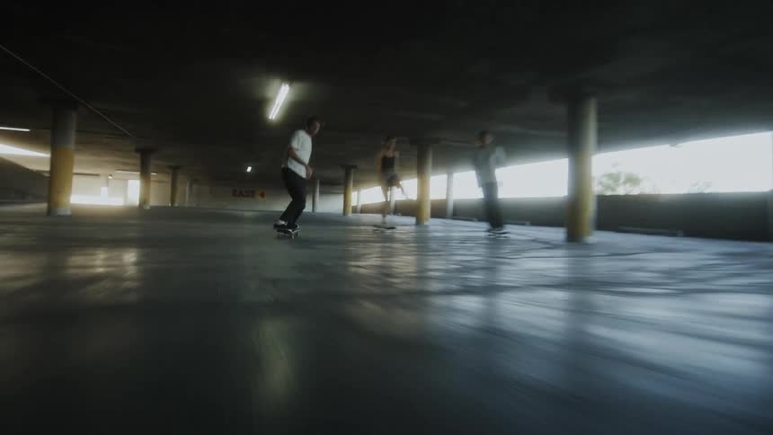 The camera rushes by three teenagers on skateboards rolling through a parking garage