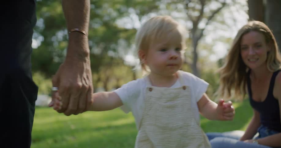 Mother watches with a smile from picnic blanket as father helps daughter toddle along