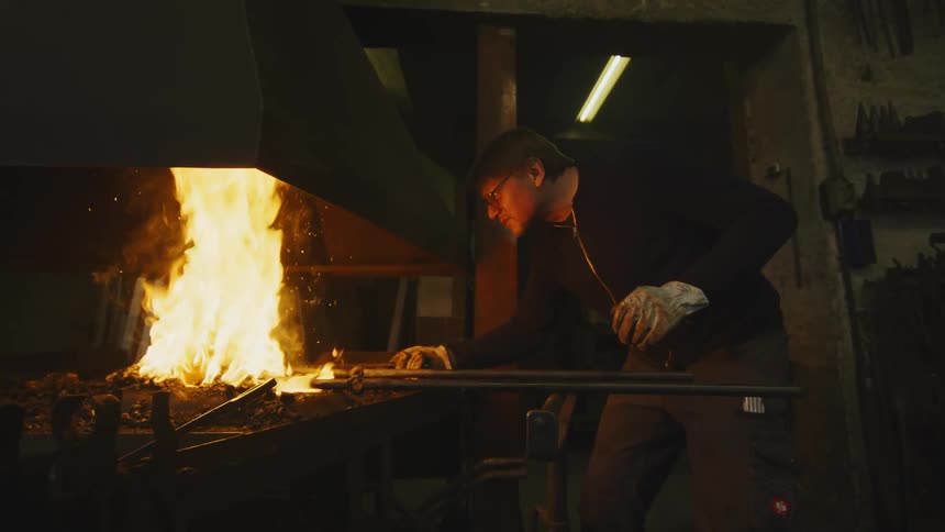 A man wearing gloves pokes at a fire with tools in a workshop