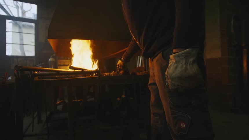 Blacksmith stands in front of the intense fire of a foundry in a dimly lit workshop