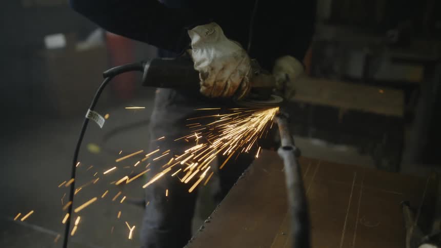 Sparks fly as a man standing in a room uses a power tool on a pipe he is holding
