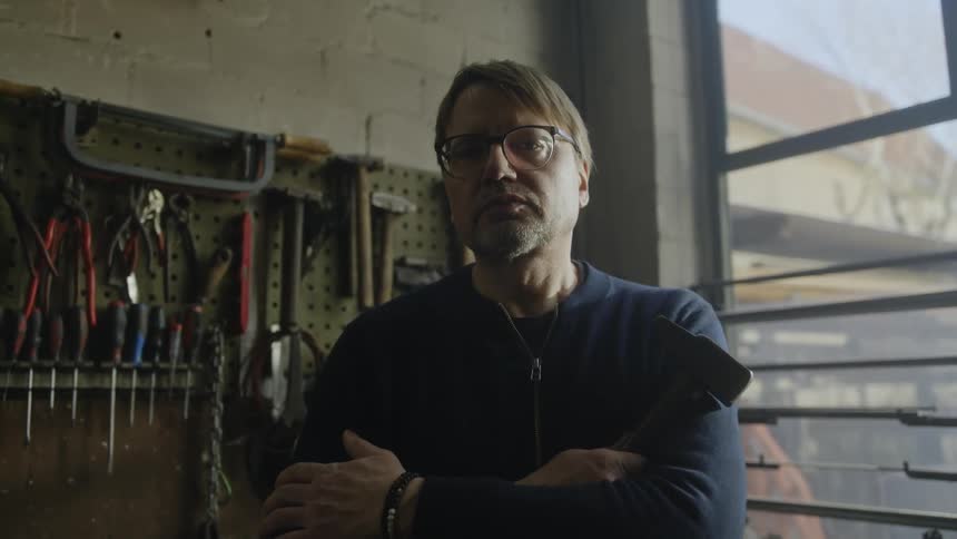A mechanic is standing in a garage and folding his arms while holding a hammer