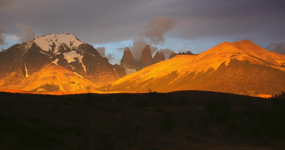 A wide mountain time lapse with clouds rolling over the peaks lit up by an orange light