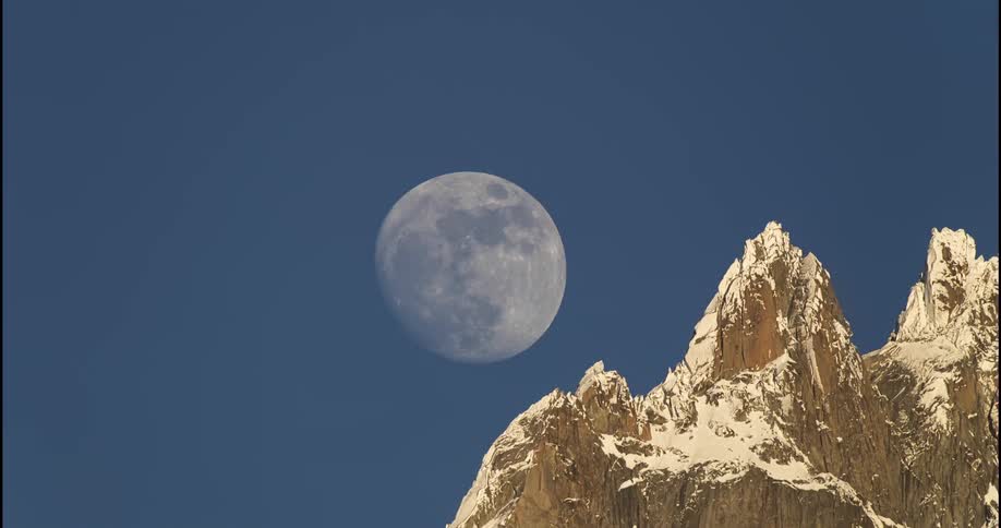 Outside during the day a full moon is shown in the sky moving slowly over some large rocks and higher into the sky