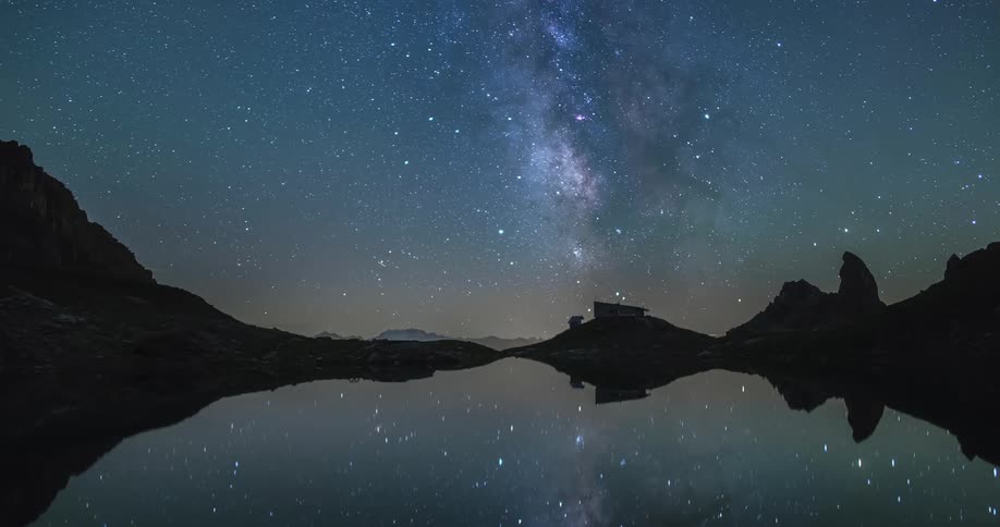 Shooting stars streak across the sky over mountains as light shines from structure near reflective lake