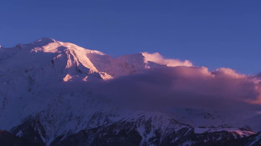 The sun shines as it sets and clouds move around snow covered mountains under a blue sky