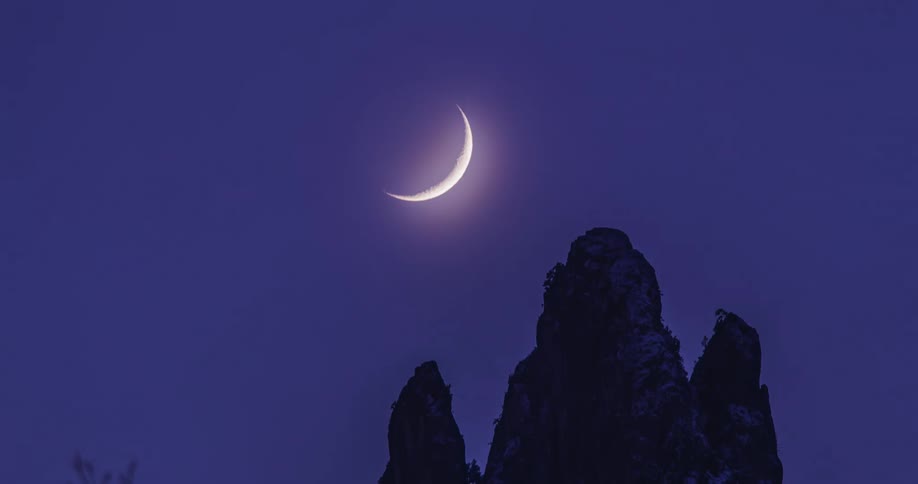 Three silhouettes of a mountains peak at night as a crescent moon moves across the night sky