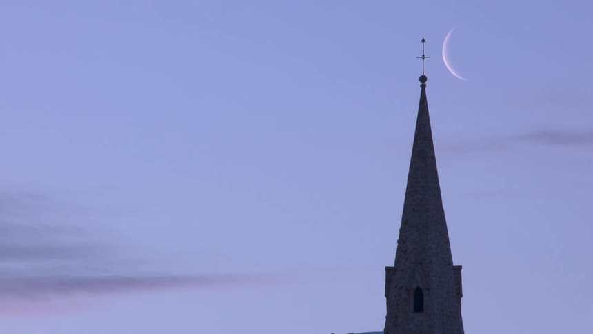 A crescent moon moves across the sky and behind a building as a bird flies from the tops of the steeple