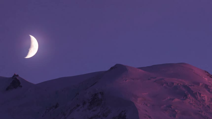 The crescent moon passes over a snowy mountain peak as the sky darkens