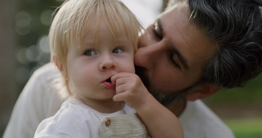 A father holds his young son outdoors and kisses his cheek