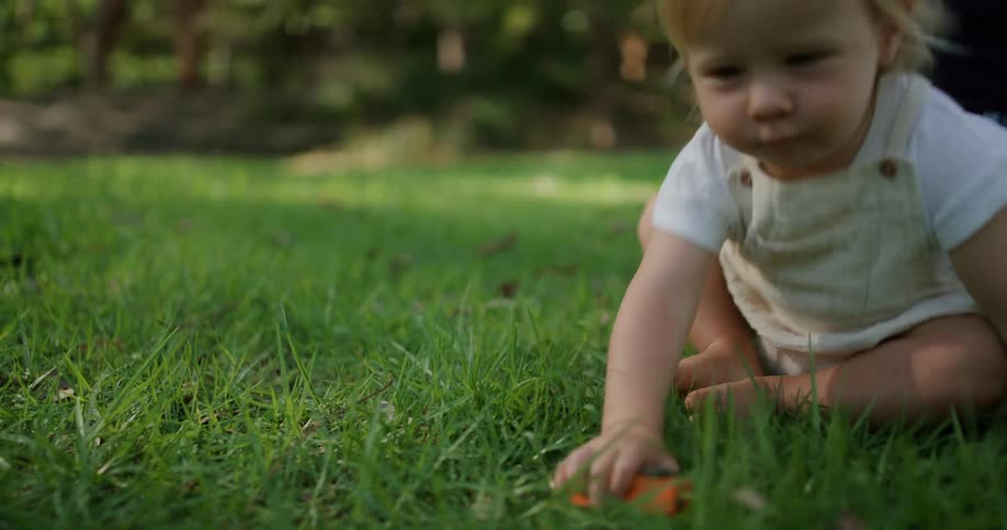 A toddler walking on grass away from an adult woman sitting on the grass behind them as the toddler falls and sits down