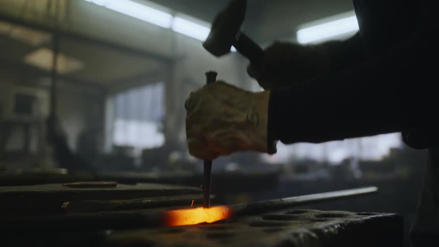A close up of a man pounding a metal stake through a hot rod with a hammer while wearing gloves