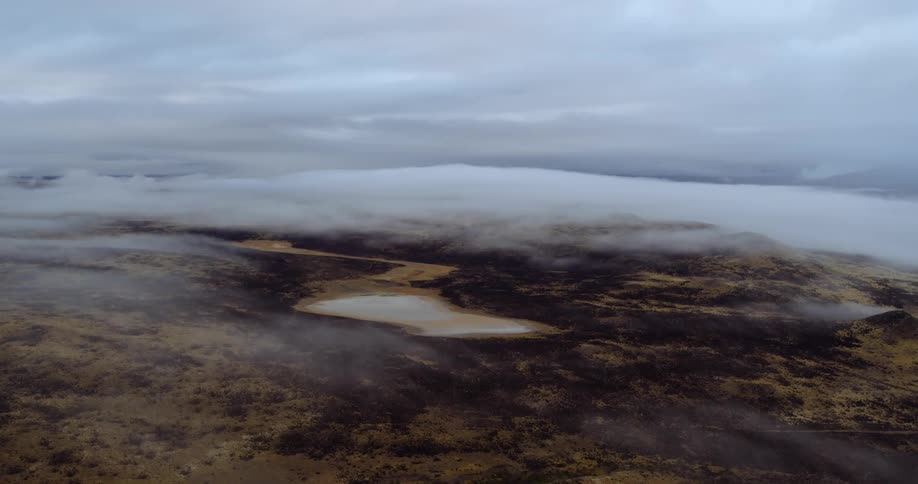 An aerial of a dark plateau of land with dark trees wispy clouds and water on the land