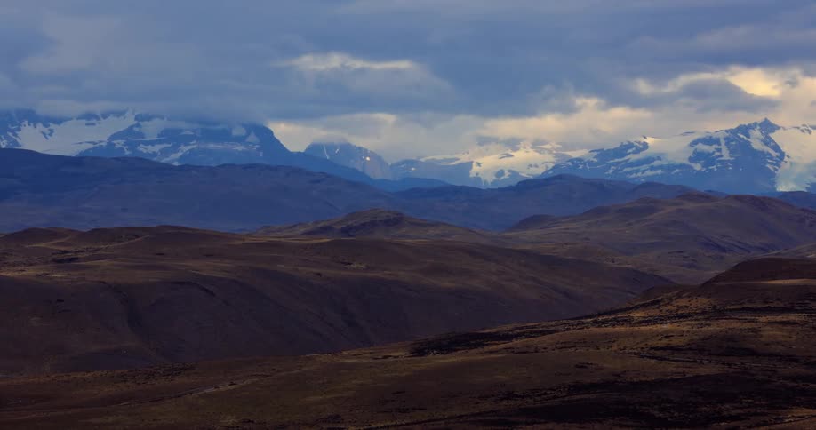Clouds cast shadows over green hills and nearby snowy mountains