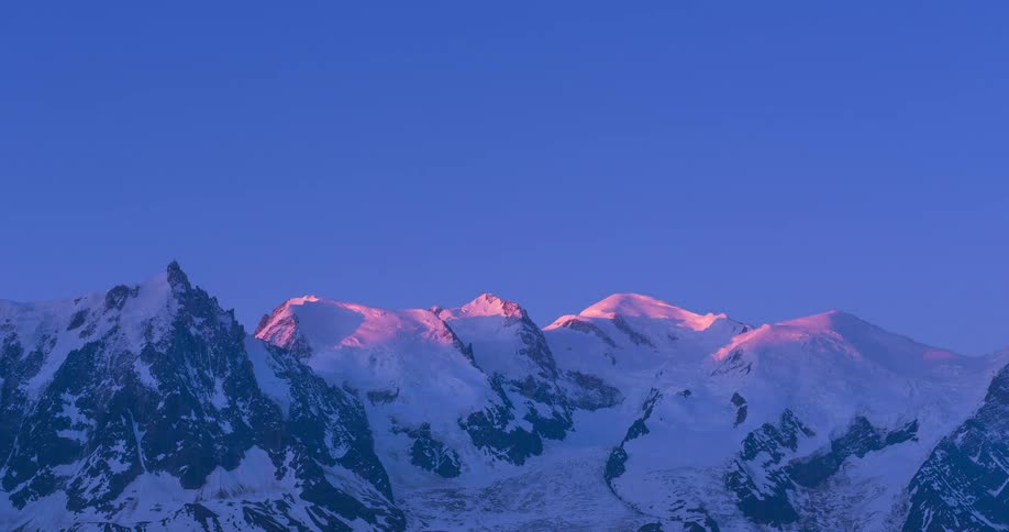 Sunlight slowly illuminates the snowy peaks of a mountain range