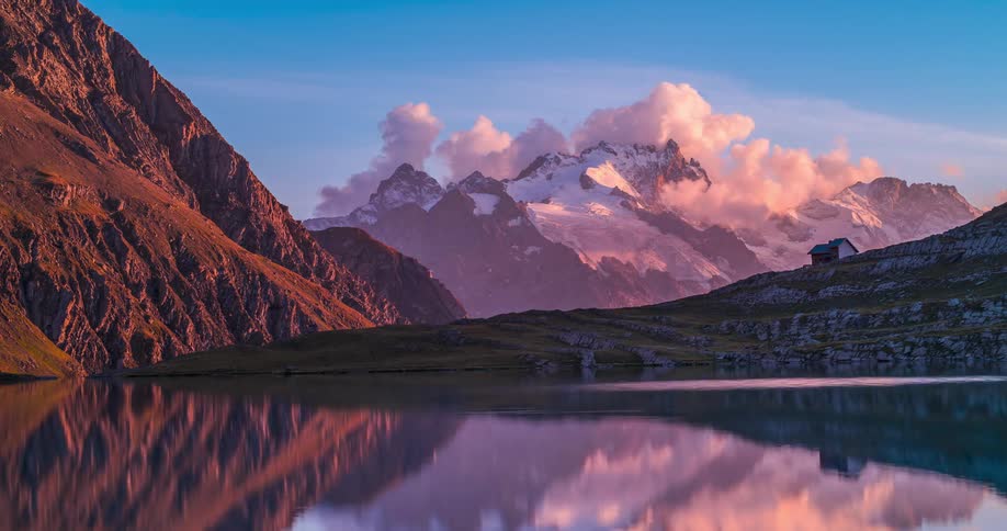 Small house sits on slope near reflective lake as clouds over nearby snowy mountain tint red during sunset then fade away