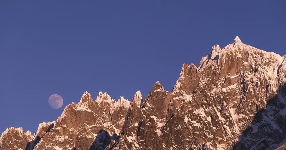 A glowing moon rises over spikey mountains as a shooting star streaks across the clear blue sky