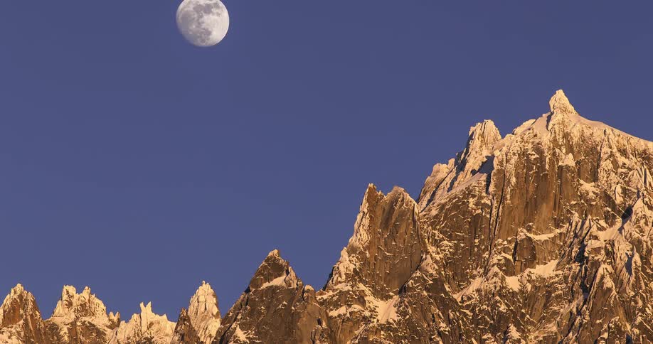 A glowing moon rises over spikey mountains in the clear blue sky