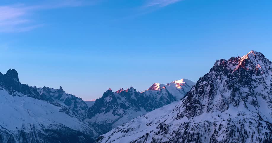 Dawn sunlight tints clouds and tops of snowy mountains while the remaining mountainsides remain shadowed
