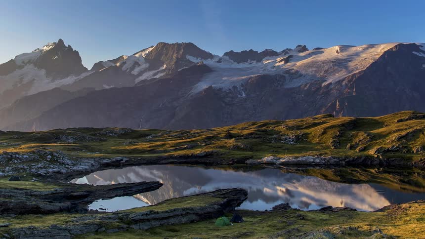 Campers move about shore around lake reflecting nearby snowy mountain as sun rises
