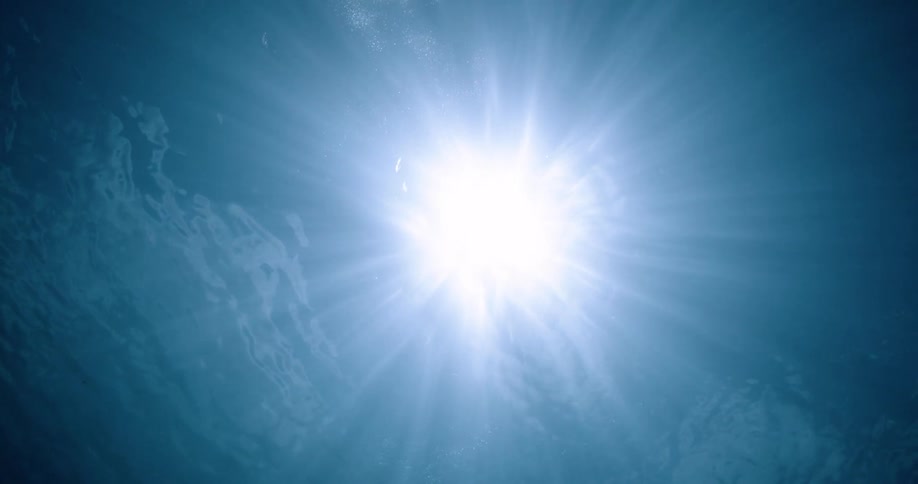 The sun and rays of light shine above as seen from beneath the surface of the water