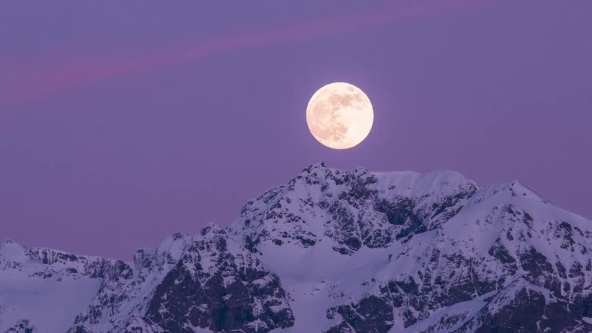 The moon rises over a snowy mountain ridge as the sky grows dark