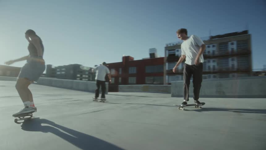 Three teenagers ride on their skateboards on a cement area with buildings in a city in the background