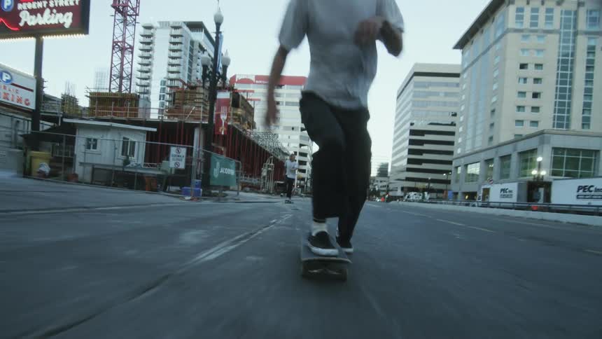 A man wearing a white t-shirt and black Vans skateboards down a city street past buildings and traffic cones