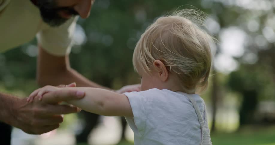 A toddler boy walking in grass towards his fahter who puts his hands out and picks up the child