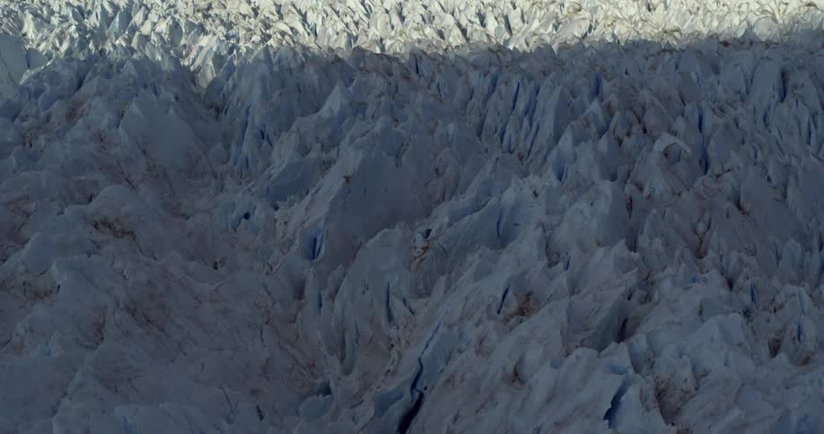 An aerial of a large ice cap with sun lit on half of it and a mountain range in the distance