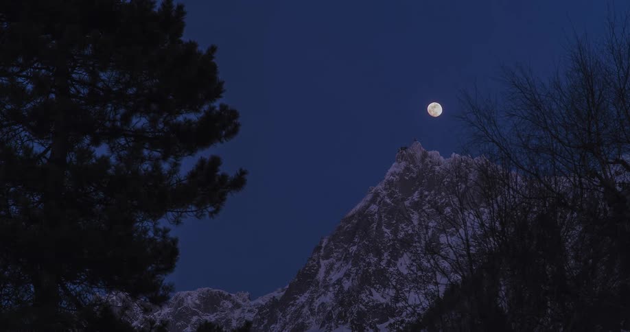 A time lapse of a tall mountain peak with a snow dusting on the ridges as the moon raises in the dark growing sky with trees in the forefront