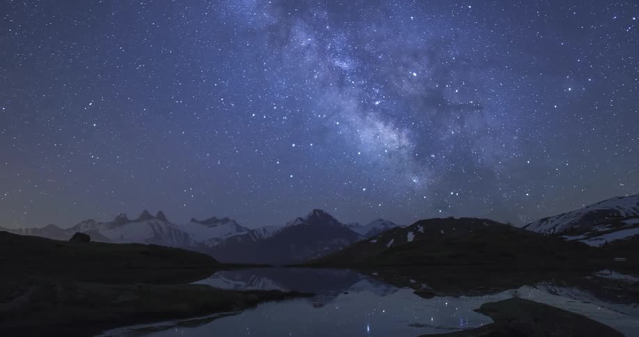 The stars of the milky way move over mountains and reflect in the water as shooting starts fly across the sky