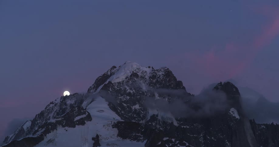 A time lapse of a full moon rising over a mountain ridge as the sky turns night and the moon reaches the peak of the snowy mountain