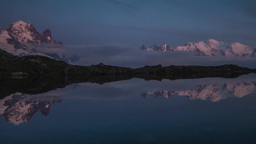 A panoramic time lapse view of snowy mountains transitioning from dusk to night while being reflected in a lake