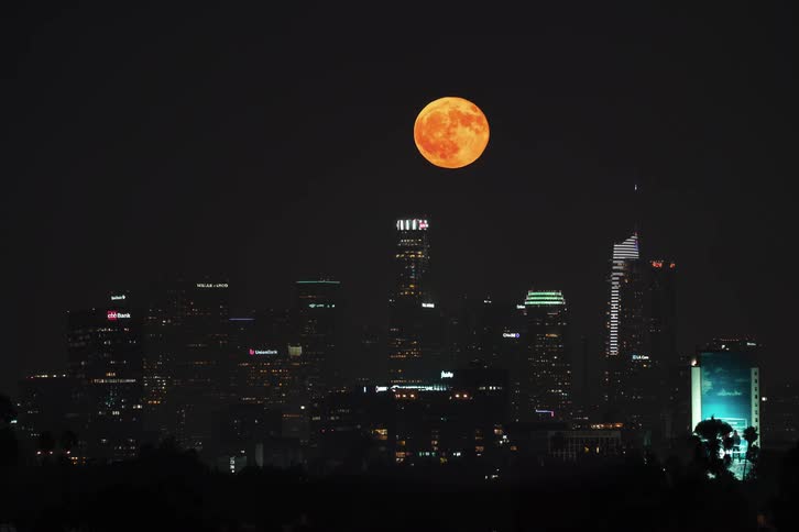 Lights on towers and skyscrapers glow in a metropolitan city as a full red blood moon rises behind the buildings and over the city in the night sky