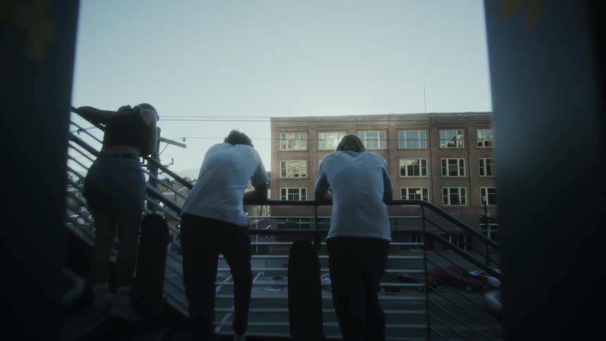 A group of three teenagers with skateboards stand near an urban staircase railing while talking