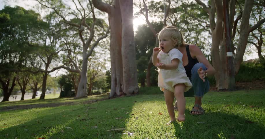 A mother crouching on grass she lets a toddler go to walk in front of her