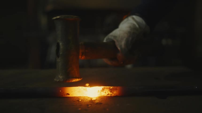 An up close view of a person wearing gloves using a sledge hammer to smash a hot steel bar
