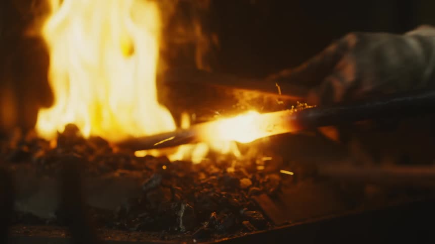 Blacksmith combs over glowing bulbous section of metal bar resting in fire