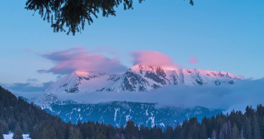 An aerial view of snow covered mountain peaks with clouds and fog rolling over the peaks and below with a forest in the valley
