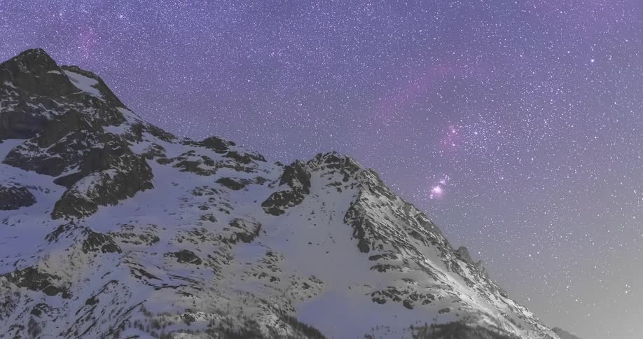 A time lapse view of the starry night sky behind a snowy mountain peak